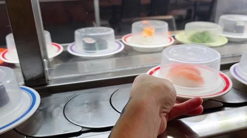 Woman's hand picks a covered sushi plate from a moving conveyor belt in a J.. Video stock 338363087