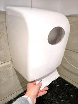 A womans hand pulls out a white paper towel from a wall-mounted dispenser. 스톡 사진