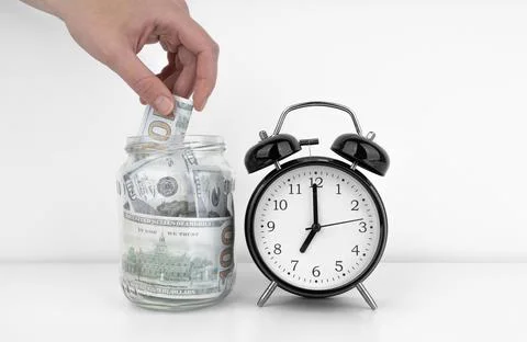 Womans hand put dollar bancnote into a glass jar with alarm clock Stock Photos