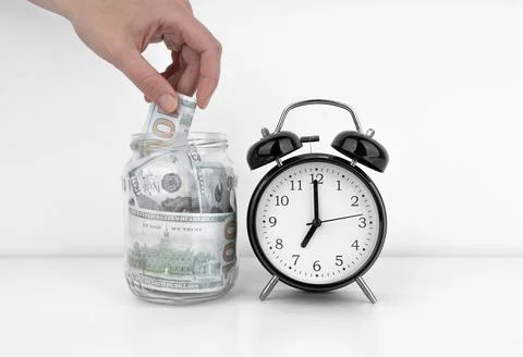 Womans hand put dollar bancnote into a glass jar with alarm clock Stock Photos