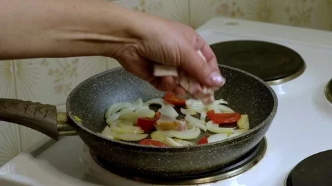 A woman's hand puts bacon in a frying pan with fried onions and tomatoes Stock Footage 242784825