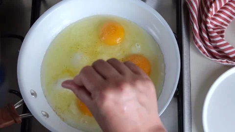 A woman's hand salts four broken eggs on a white frying pan. in the kitchen top Stock Footage 254546426