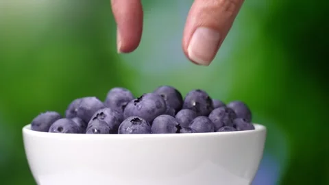Woman's hand takes and puts berry of ripe selected blueberries from plate Stock Footage 248783169