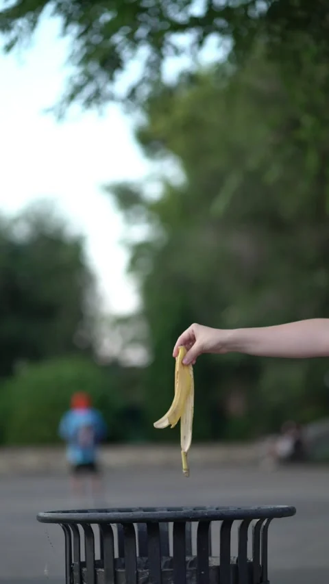 A woman's hand throws a banana peel into a metal trash bin in a park Stock-Footage 340670487