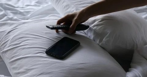 Woman's hand using TV remote control and smartphone on bed in the morning Video stock 112242787