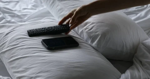 Woman's hand using TV remote control and smartphone on bed in the morning Video stock 112243009
