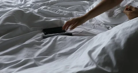 Woman's hand using TV remote control and smartphone on bed in the morning Stock Footage 112243786