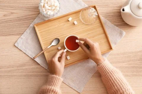Womans hands adding sugar cube to a cup of hot tea on a wooden tray, top view Stock Photos