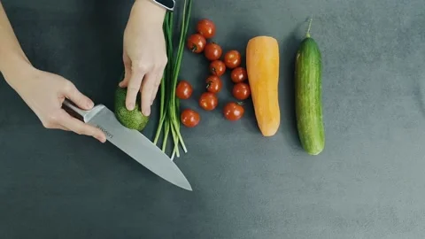 Woman's hands cuts avocado on two halves on the table with one fresh cucumber Stock Footage 136114083