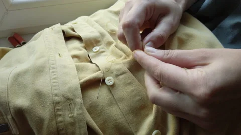 Woman's hands getting ready to sew button to yellow shirt. Female at work Stock Footage 137276702