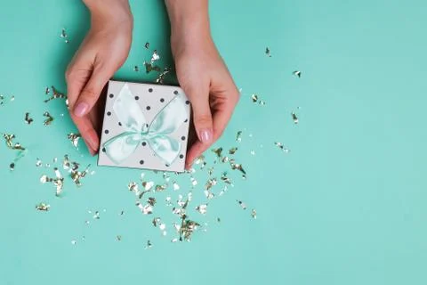 Woman's hands holding a gift box on on turquoise background with golden confetti Stock Photos