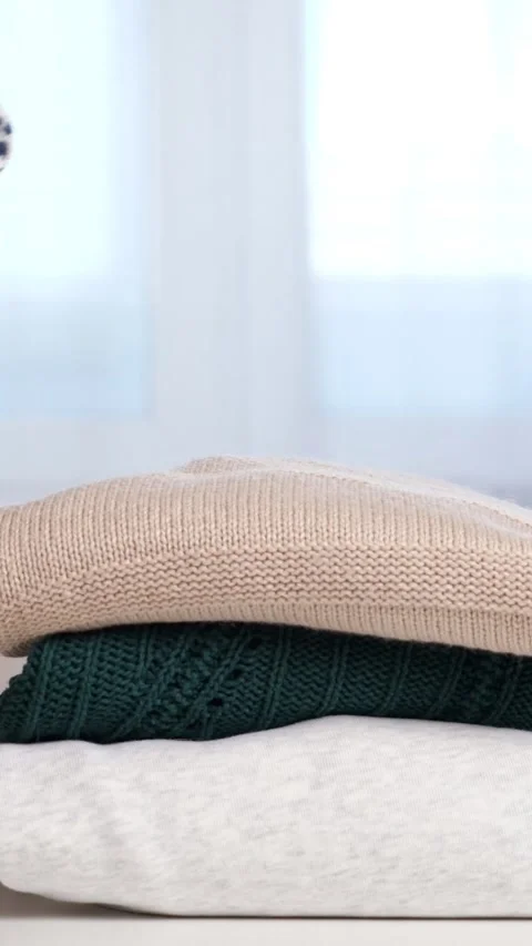 Womans hands neatly folding warm knitted sweaters into a tall stack on a white Stock Footage 343910286