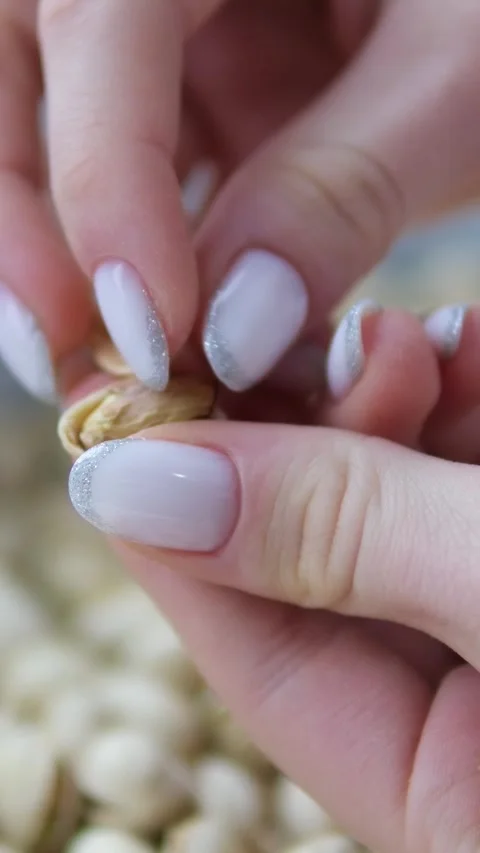 Woman's hands opening a pistachio nut on a table surrounded by more pistachios Stock Footage 302106492