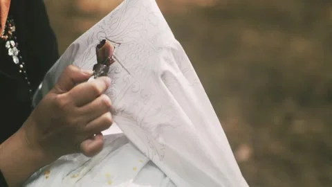 A woman's right hand holds a canting tool for batik on a white cloth. 스톡 동영상 329594765