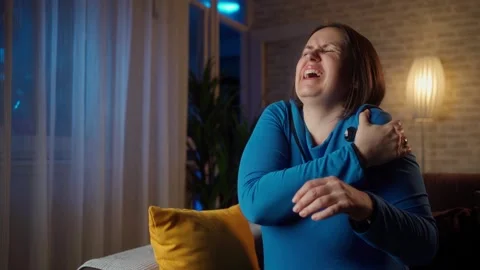 Woman's shoulder hurts while sitting on the sofa at home in the evening Stock Footage 268327536