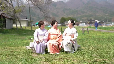 Women in kimonos chat while seated on a spring lawn Stock Footage 344793442