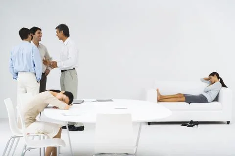 Women napping, one at table, one on couch, men talking in group in the corner Stock Photos