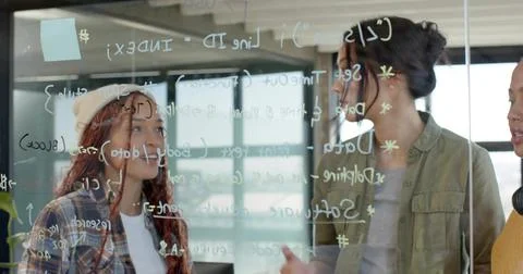 Women in varied attire collaborating on code behind glass board in office, wi Foto stock