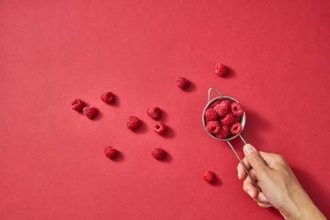 A womens hand holds a colander with red ripe sweet raspberry on a red paper with Foto stock
