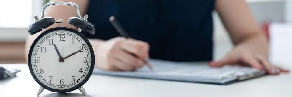 Women's hands write in the document. On the table is an alarm clock. Stock Photos