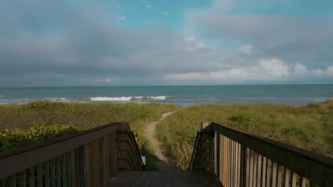 A wooden boardwalk ends at a beach access path, framed by grassy dunes and a Stock Footage 312725698