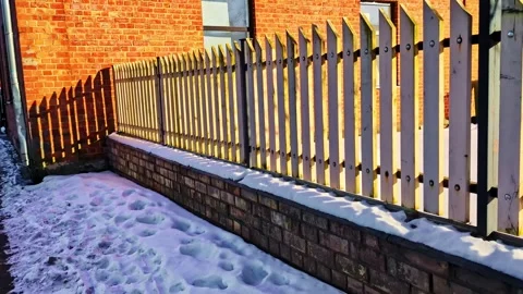 Wooden fence casting long shadows on a brick wall and snow-covered ground Stock Footage 332744155