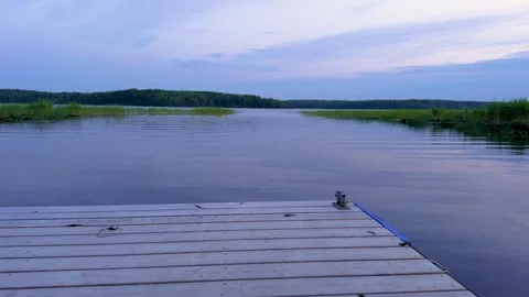 Wooden pier extends into a tranquil lake at dusk with a forested shoreline Stock Footage 316133653