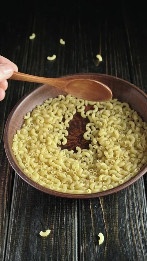Wooden spoon scooping elbow macaroni from a brown bowl on a dark wooden table Stock-Footage 338412239