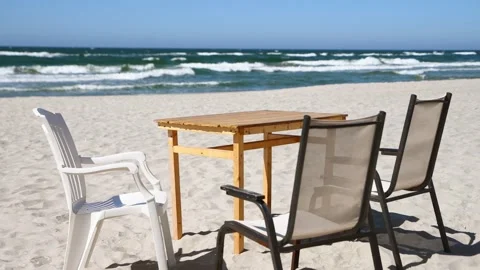 Wooden table with plastic and mesh chairs on sandy beach by blue sea under .. Stock Footage 335941979