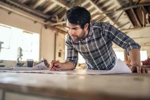 Woodworker leaning on a workbench in his studio reading blueprints Stock Photos