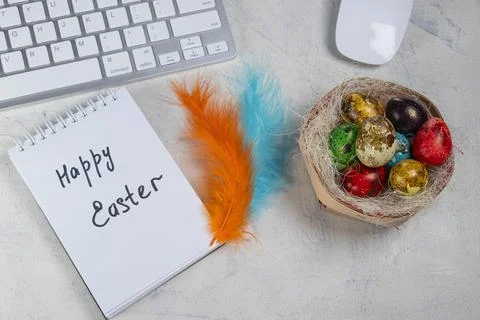 Words Happy Easter in a notebook on the desktop with colored eggs in the nest Stock Photos
