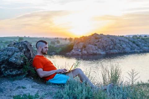 Workaholic working on a laptop while sitting on a stone, beautiful sunset. Stock Photos