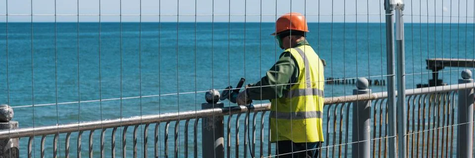 Worker with angle grinder. Work on the restoration of the embankment fence. 2 Stock Photos