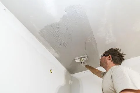 Worker applying joint compound to ceiling with trowel during interior renovation Stock Photos