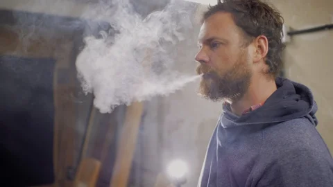 Worker with a beard and mustache smokes a cigarette indoors against a backlight Stock Footage 105941156