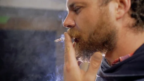 Worker with a beard and mustache smokes a cigarette indoors against a backlight Stock Footage 105941255