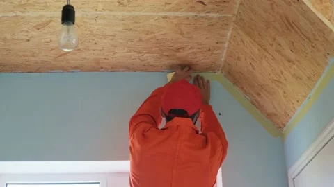A worker with beard in an orange robe is sticking painter's tape on the wall bef Stock Footage 197785745