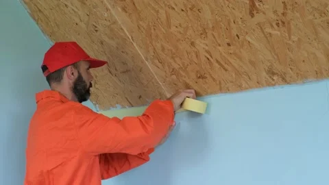 A worker with beard in an orange robe is sticking painter's tape on the wall bef Stock Footage 197785794