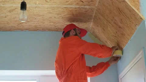 A worker with beard in an orange robe is sticking painter's tape on the wall bef Stock Footage 197785808