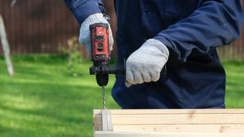 A worker in a blue uniform and protective gloves drills a hole in a wooden .. Stock Photos