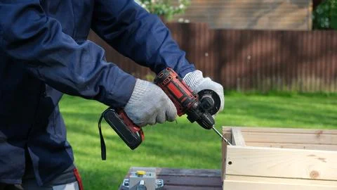 A worker in a blue uniform and protective gloves drills a hole in a wooden .. Stock Photos
