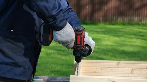 A worker in a blue uniform and protective gloves drills a hole in a wooden st Stock Photos