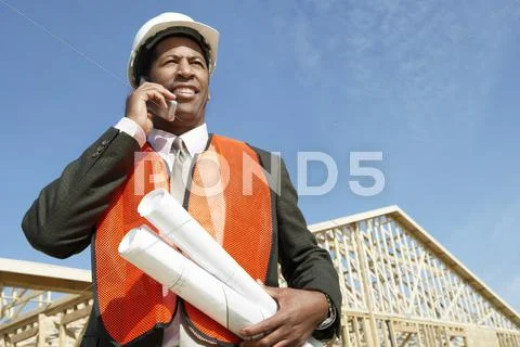 Worker With Blueprints Using Cell Phone At Construction Site Stock Photos
