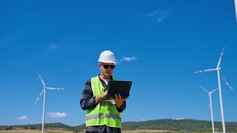 Worker checks data at wind farm on a sunny day. A person wearing a hard hat and Stock Footage 342737506