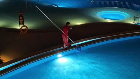 Worker cleaning illuminated swimming pool at night using long pole net Stock Footage 331584847