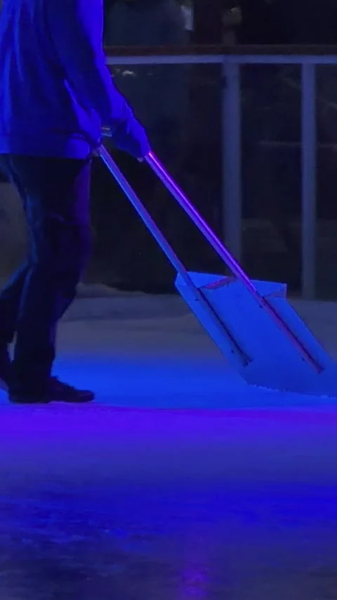 A worker cleans an ice rink surface with a large manual scraper tool under blue Stock Footage 345665679