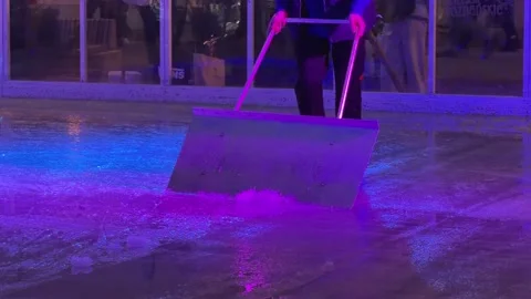 A worker clears slush from a glowing ice rink with a large metal scraper during Stock Footage 345665930
