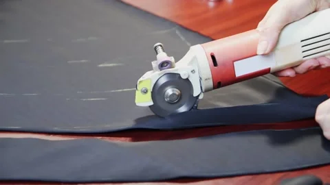 Worker cuts layers of dark tissue with electric cutter on a brown table. FullHD Vídeo Stock 141172381