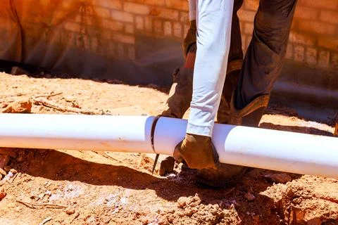 Worker cuts white pipe with tool while standing on dirt in construction area 스톡 사진