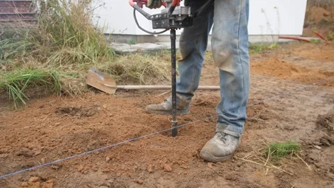 Worker drills post, laborer images onsite drilling, worker uses auger in muddy Stock Footage 319402769